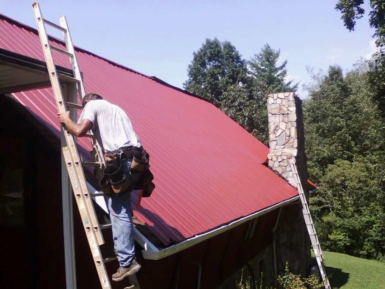 Roofer on a ladder beside a red metal roof and stone chimney — Soffit & Fascia Repair by Roofing Brothers in Gordonsville